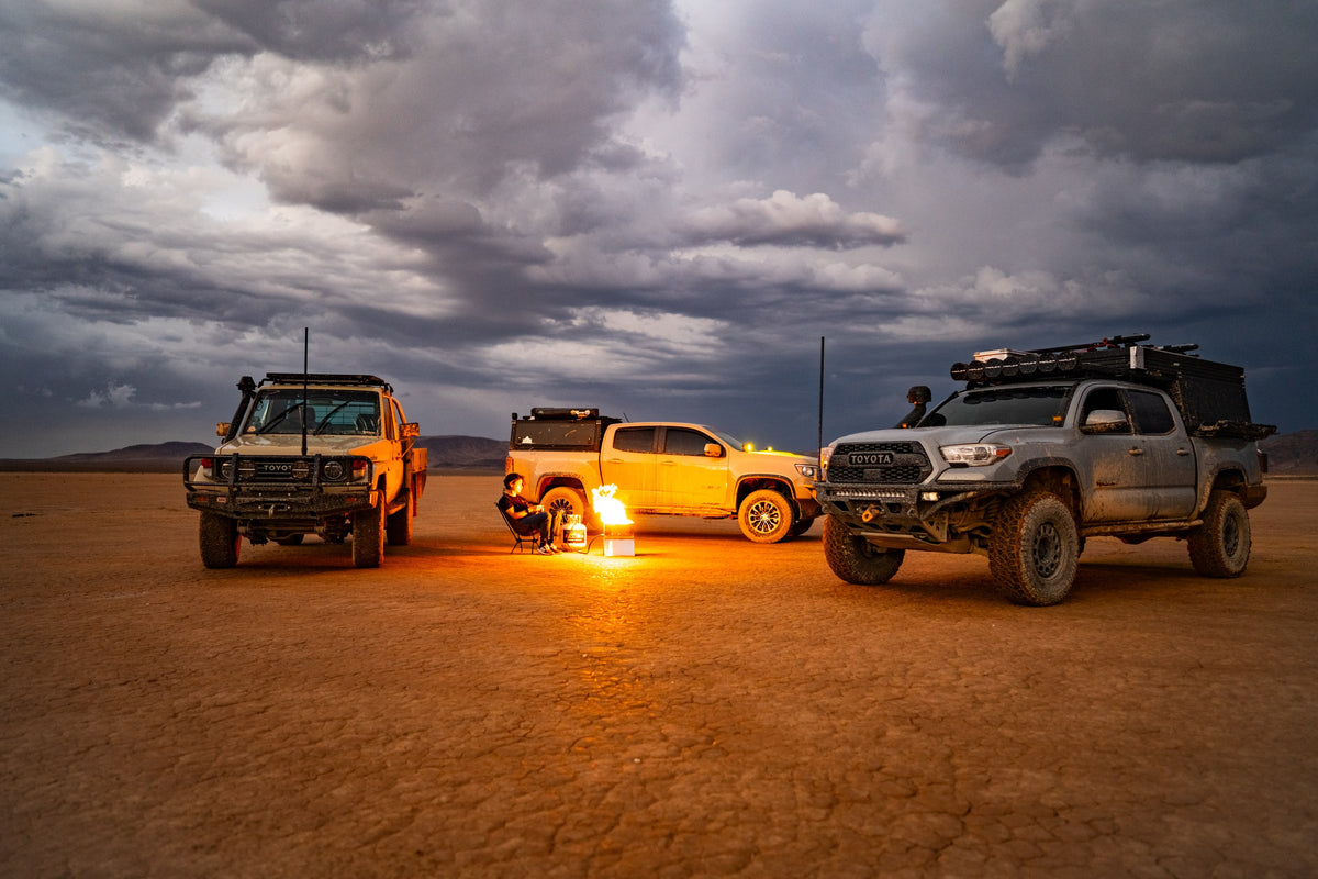 Three overland trucks parked around an RTE FireFold fire pit at dusk